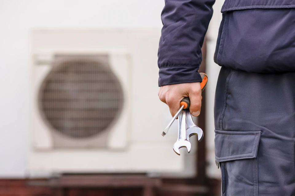 Un technicien de l'ACC portant un chapeau et un pantalon vert travaille assidûment sur un climatiseur ou une pompe à chaleur à l'extérieur