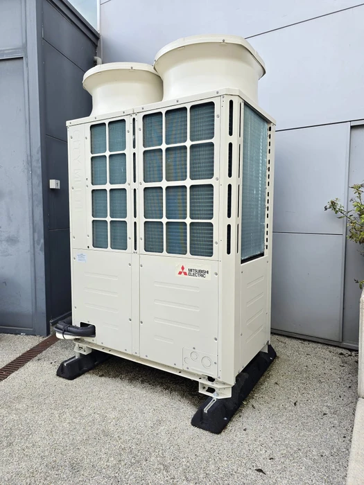 Unité extérieure de climatisation Mitsubishi Electric installée contre un mur crépi beige d'habitation, avec fenêtres visibles en arrière-plan